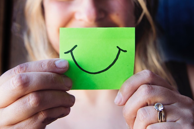 Woman holding a smiley sticky note in front of her face.